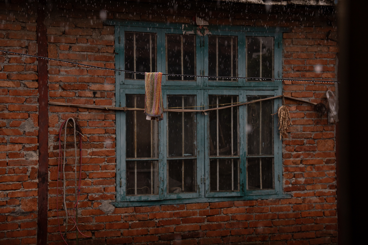 Rain falling in front of a teal wooden window in a brick wall, with a towel hanging on a line.