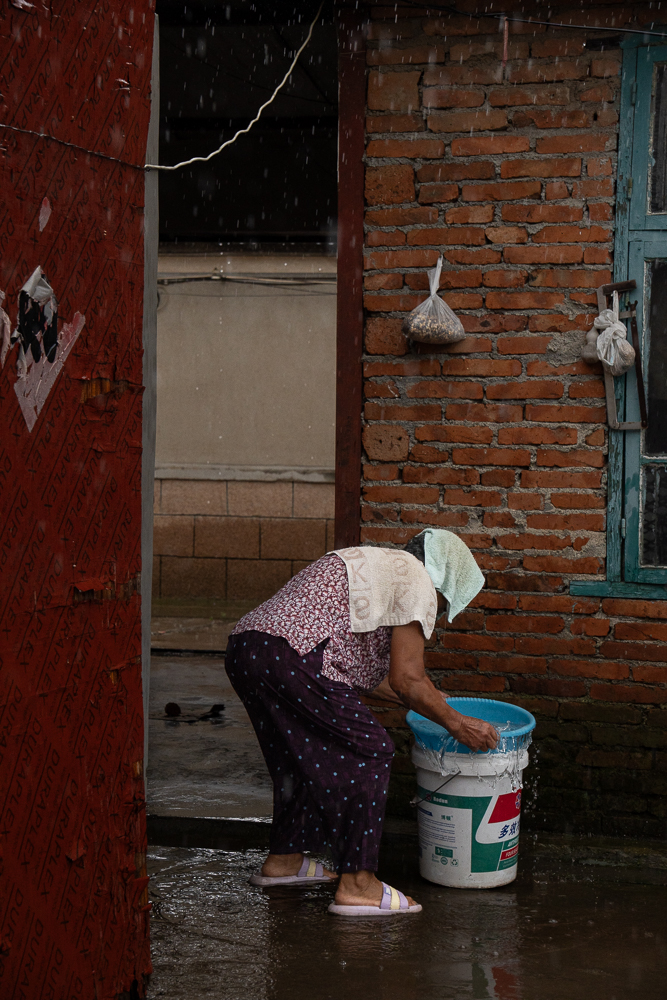 A woman bending over a bucket in a rain-soaked brick courtyard.