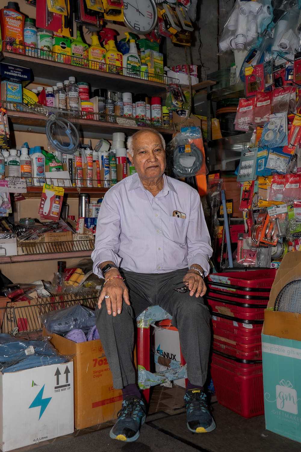 An older shopkeeper seated among hardware supplies, stacked baskets and boxes beneath a wall clock.