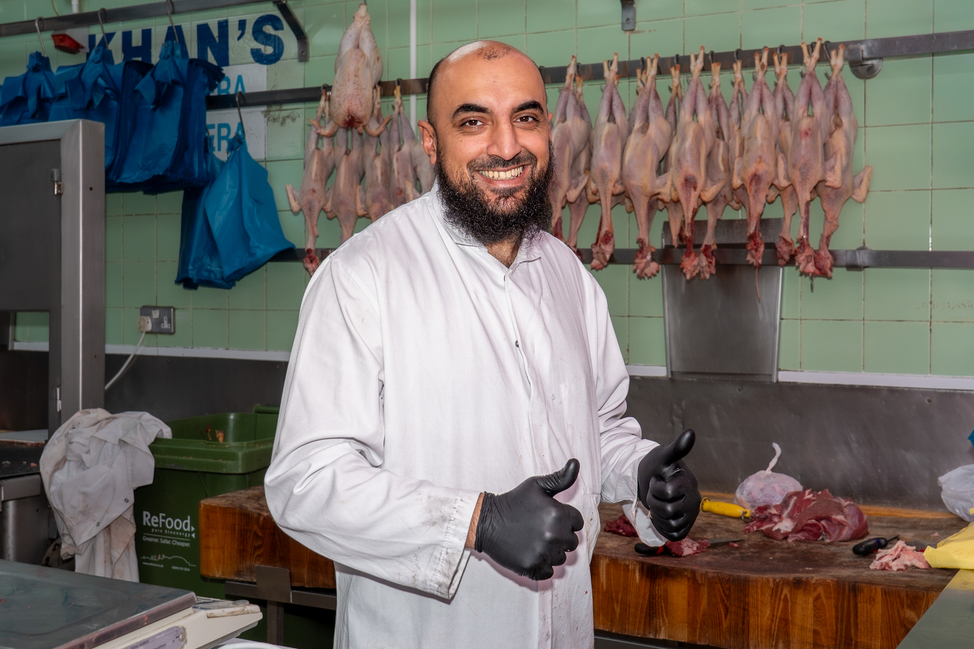 A smiling butcher in a white coat and black gloves, standing by a wooden chopping block with poultry hanging behind him.