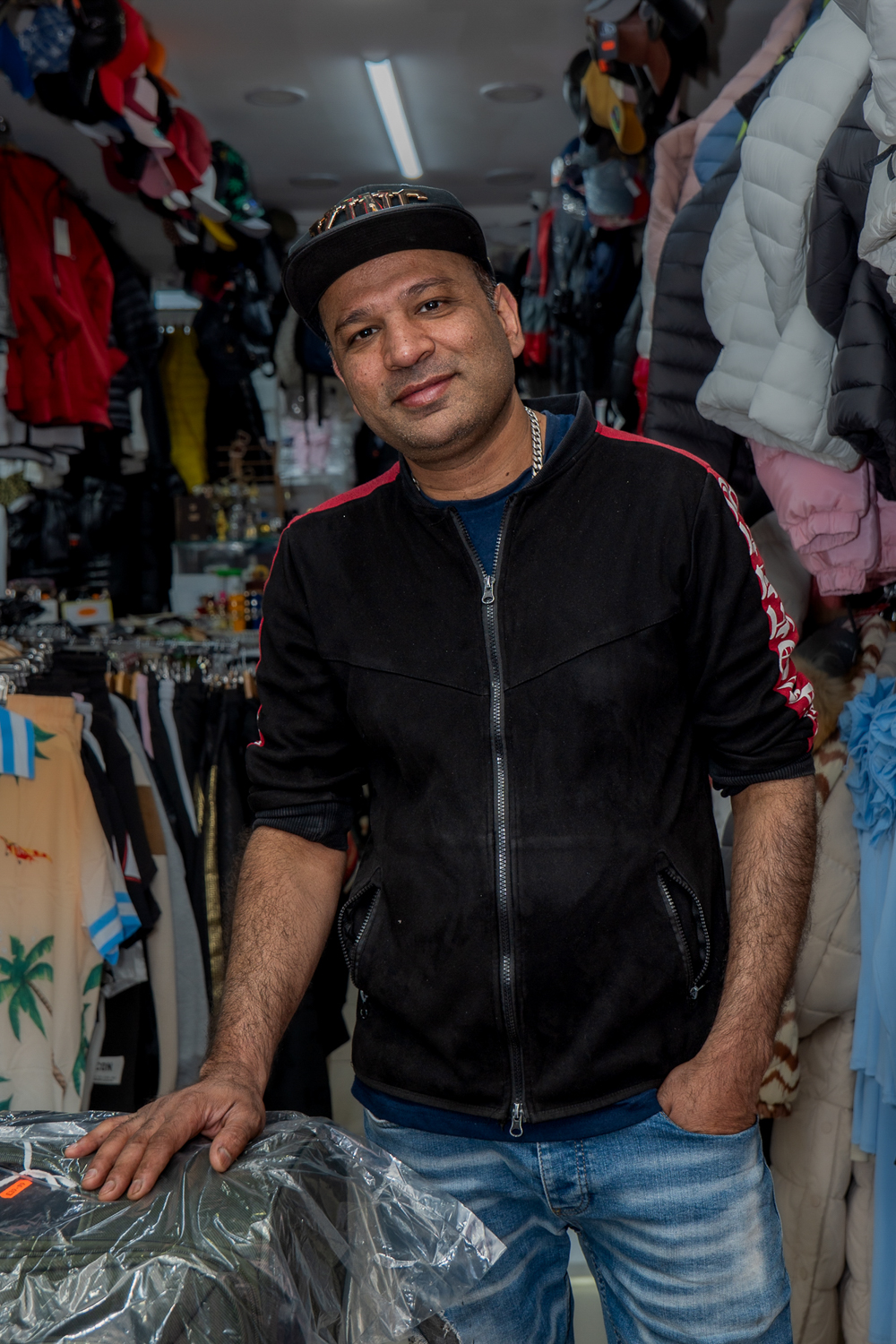 A shopkeeper standing at the counter of a narrow clothing shop lined with jackets and hats.