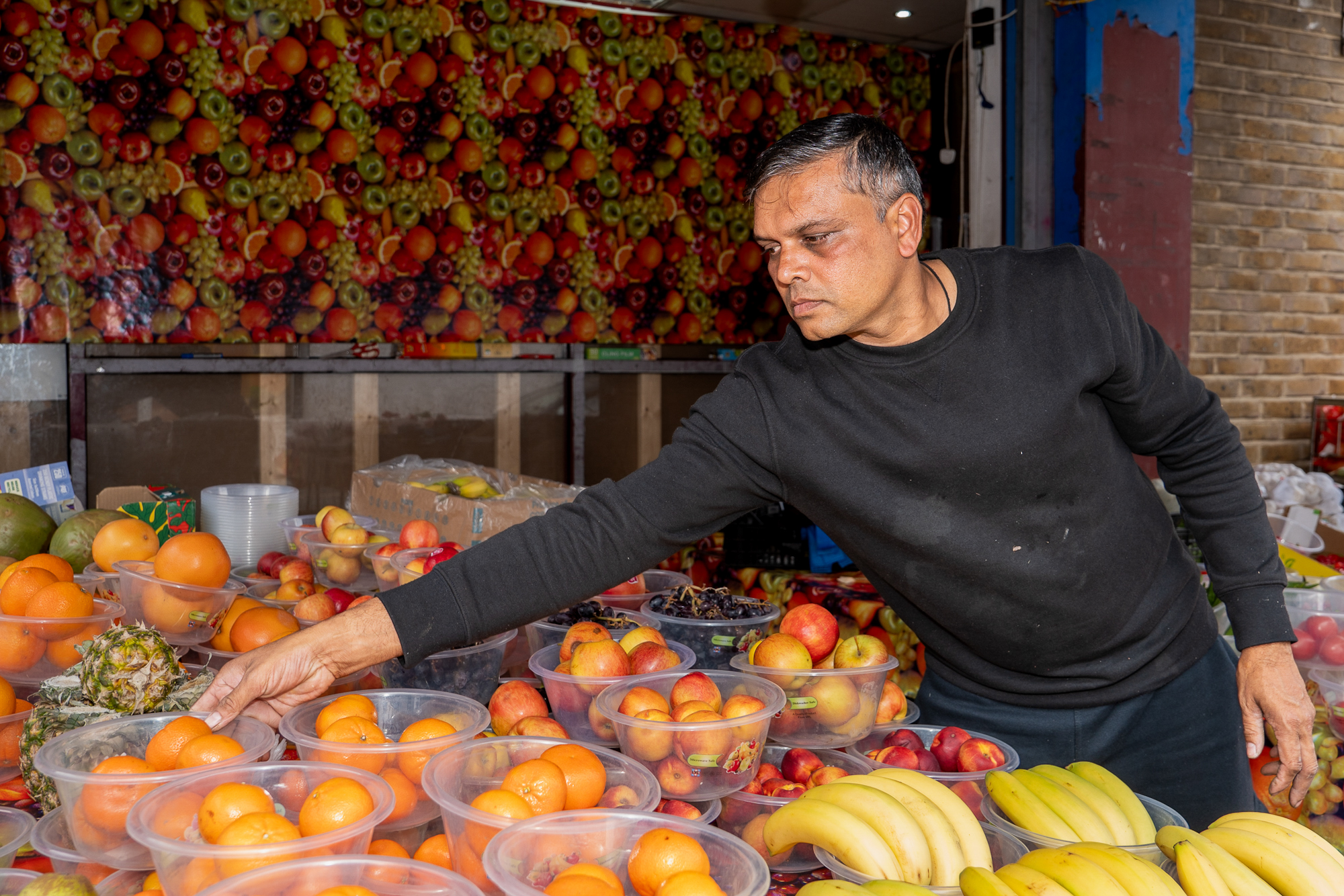 A shopkeeper reaching across a display of fruit outside a shop with a colourful fruit-patterned wall.