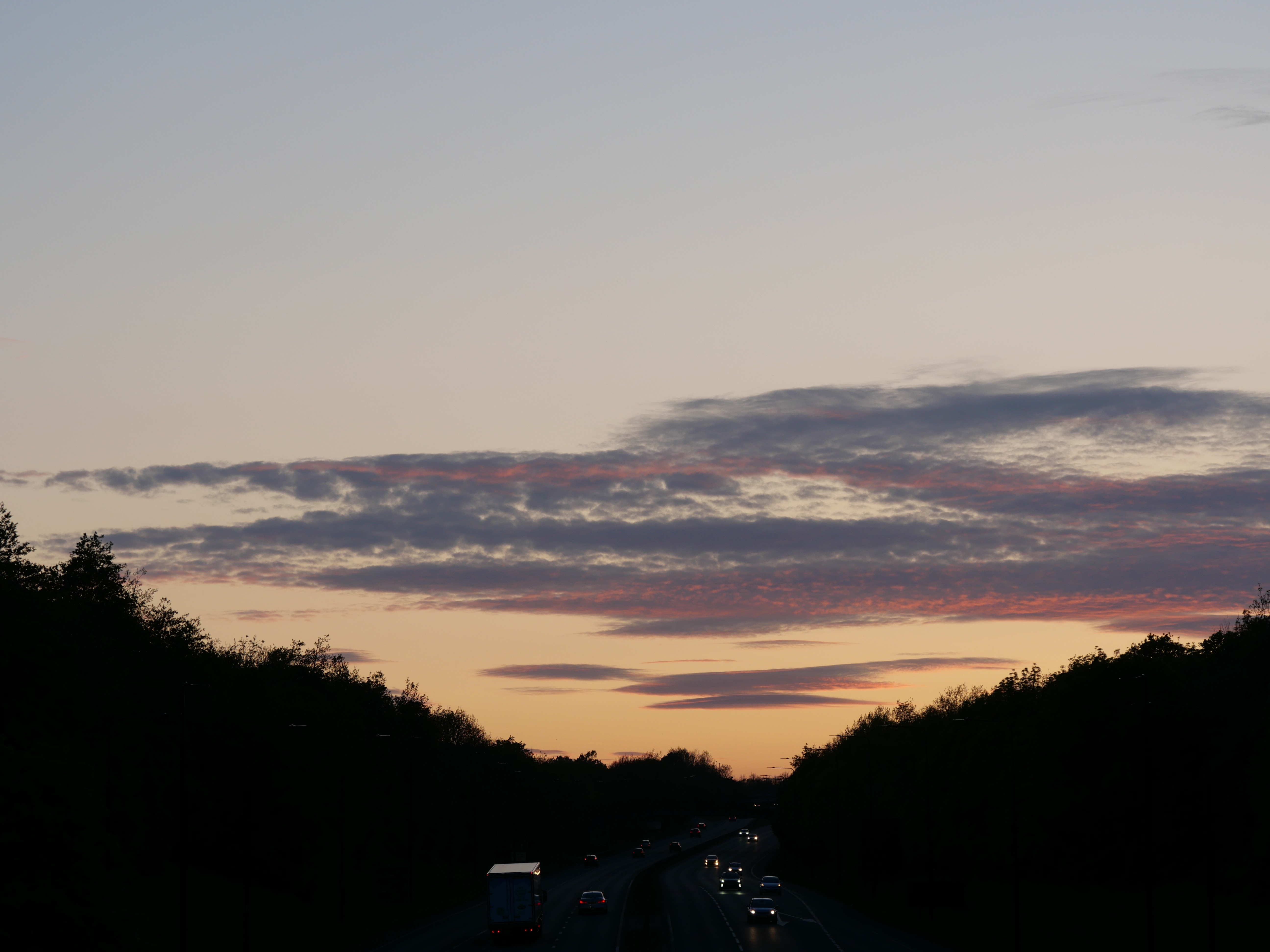 Pink and violet sunset clouds above a road bordered by dark trees.